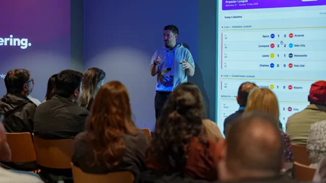 Ed Marshall presenting ScoutCast, the live football stats app, with match scores on the big screen as the audience watches